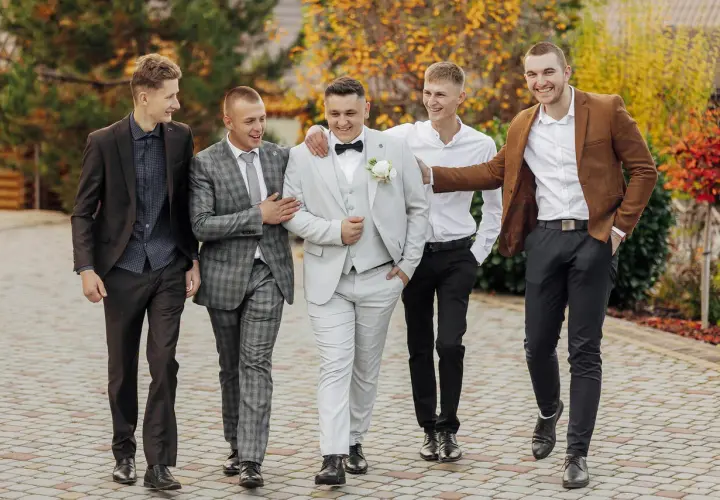 Pięciu mężczyzn w eleganckich garniturach idzie razem. Inspiracja, jak ubrać się na wesele mężczyzna.