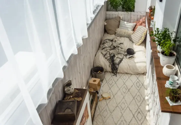 Przytulny balkon w bloku z materacem, kocem i poduszkami. Dywanik i rośliny dodają uroku.