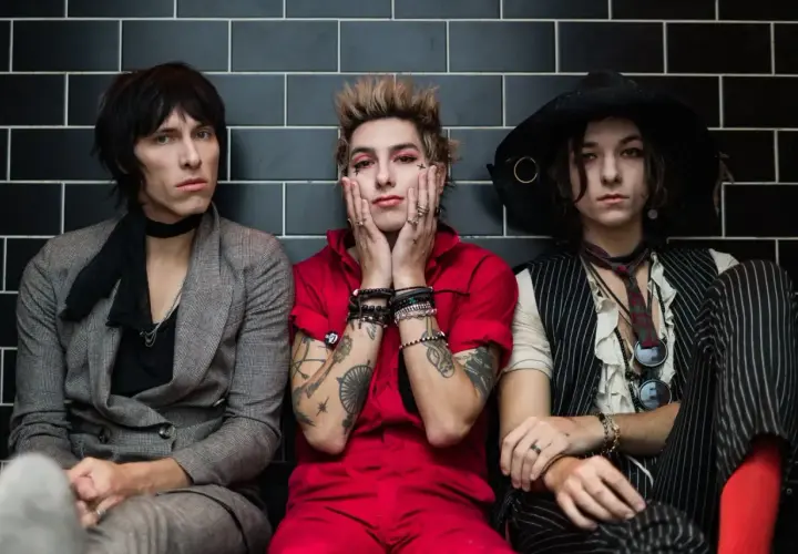 Trzech członków Palaye Royale w stylowych, rockowych strojach siedzi obok siebie na tle czarnych płytek.
