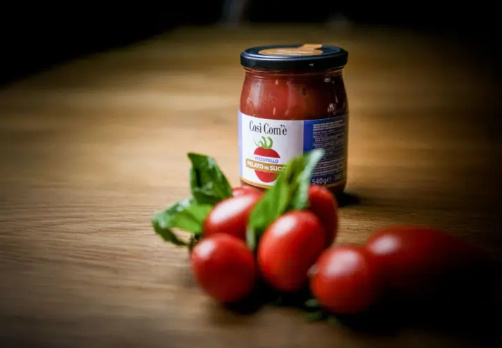 Barattolo di pomodoro siccagno pizzutello pelato in succo, con pomodorini freschi e basilico su un tavolo di legno.