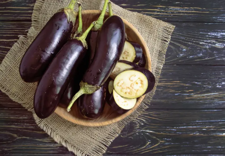 Einige ganze Auberginen liegen neben geschnittenen Scheiben in einer Holzschale. Man sieht das helle Fruchtfleisch der Aubergine innen.