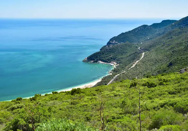 Costa verde en el sur de Lisboa, con mar turquesa, playa, barcos y montañas cubiertas de vegetación.