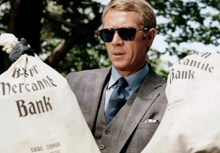 Steve McQueen w okularach przeciwsłonecznych, w eleganckim garniturze, trzyma worki z napisem "Mercantile Bank". Scena z filmu "Afera Thomasa Crowna".