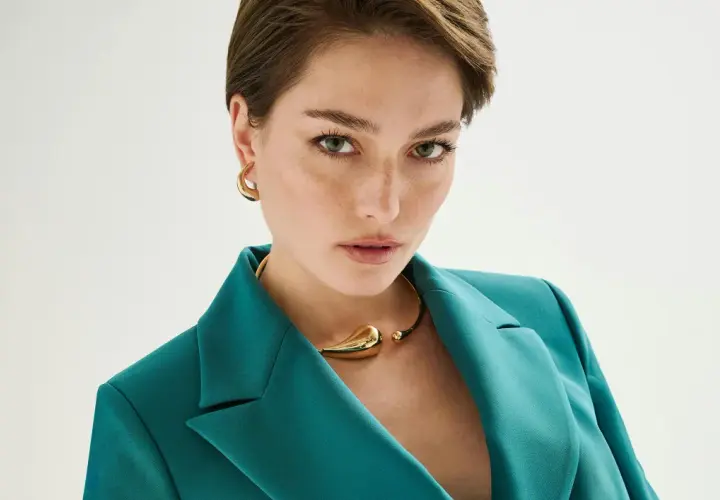 Mujer con un elegante corte pixie, viste un blazer verde azulado y un collar dorado.