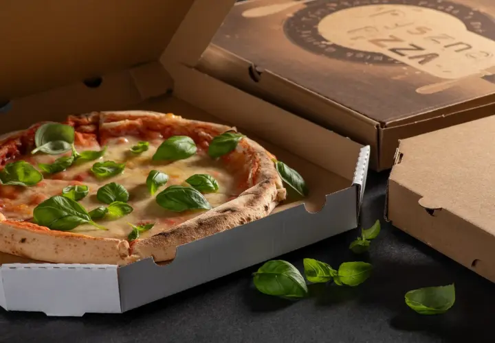 Pyszna pizza w otwartym opakowaniu na pizze, ozdobiona świeżą bazylią. Obok zamknięte kartony.