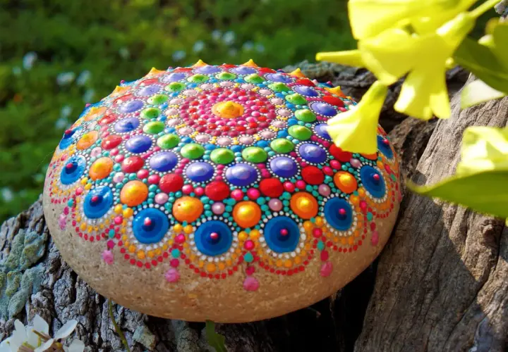 Bunte bemalte Steine mit Mandala-Muster, die auf einem Baumstamm liegen, umgeben von grünem Gras und gelben Blüten.