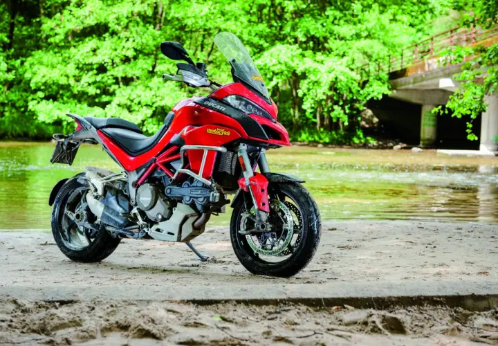 Czerwony Ducati Multistrada 1200S zaparkowany na piaszczystym brzegu rzeki, otoczony bujną zielenią.