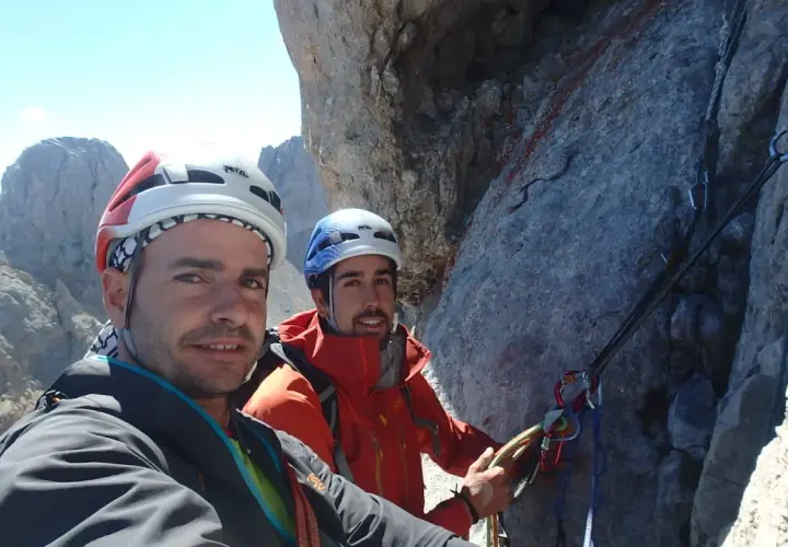 Dos escaladores sonríen en la cara del Naranjo de Bulnes, equipados con cascos y arneses, listos para la aventura.