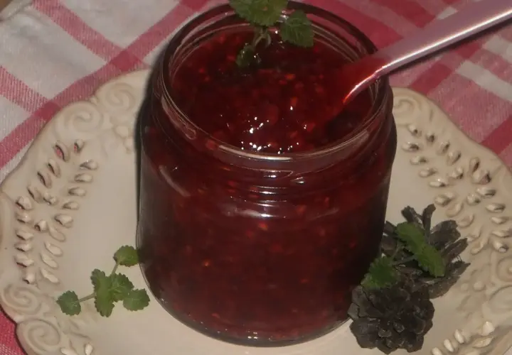 Słoik domowej frużeliny malinowej, z łyżeczką zanurzoną w gęstym, czerwonym musie. Udekorowana listkami mięty i szyszkami.