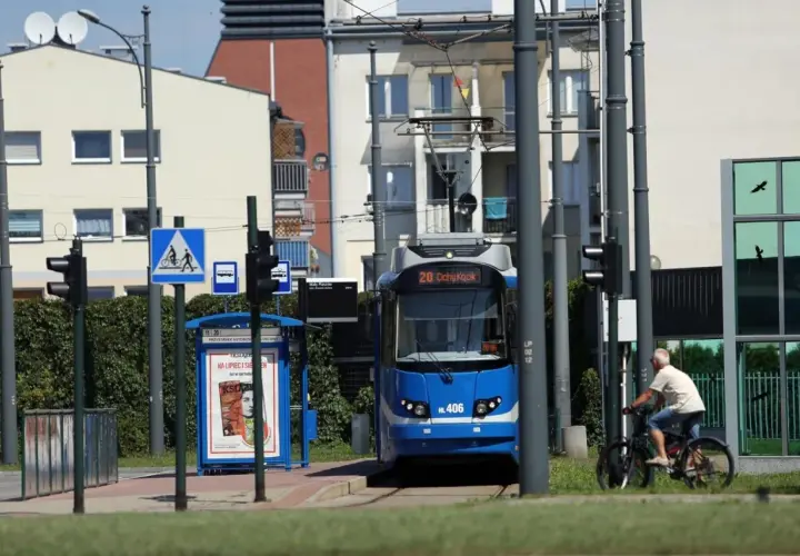 Niebieski tramwaj linii 20 na przystanku, obok rowerzysta i budynki.