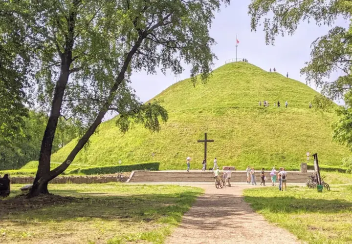 Zielony Kopiec Piłsudskiego z krzyżem i ludźmi spacerującymi po zboczu.