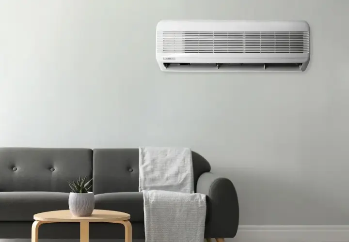 Unidad de aire acondicionado montada en la pared, cumpliendo la normativa aire acondicionado. Sofá gris con mantas y mesa auxiliar con planta.