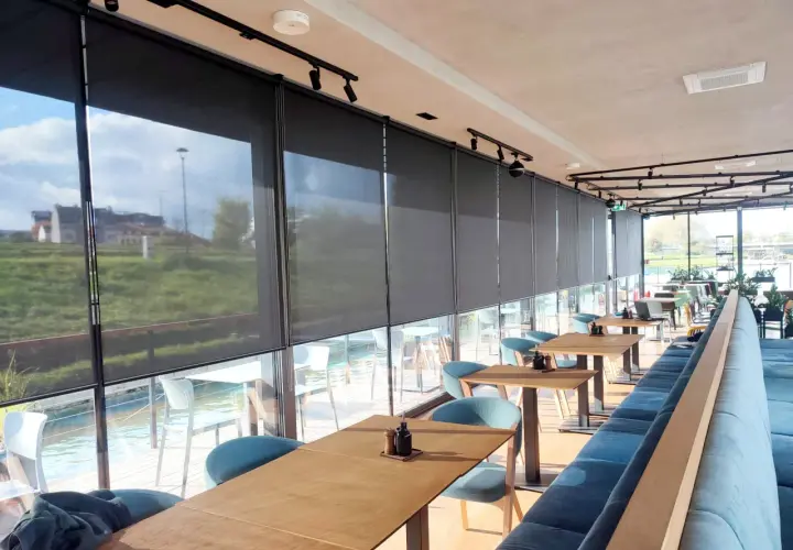 Nowoczesne wnętrze restauracji z widokiem na wodę, osłonięte eleganckimi roletami termicznymi na wymiar.