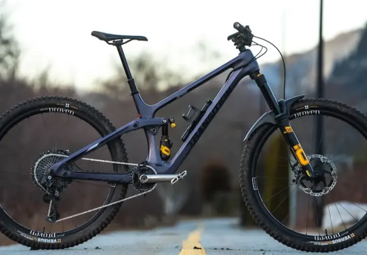 A dark purple full-suspension mountain bike with yellow accents sits on a road. Its components suggest a focus on performance, hinting at a competitive mountain bike weight for agile trail riding.