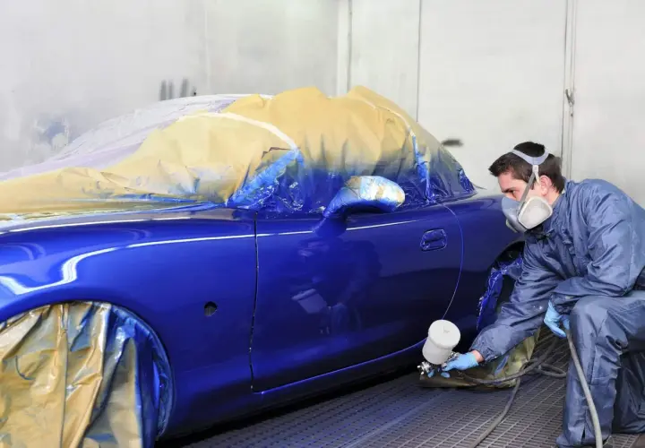 Un carrozziere con maschera spruzza vernice blu su un'auto coperta. Consulta il listino prezzi verniciatura auto per i costi.