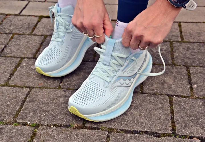 Dłonie zawiązują sznurówki jasnoniebieskich butów do biegania Saucony Ride 18 na chodniku z kostki brukowej.