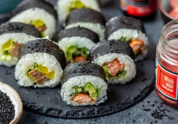 Gotowe do jedzenia rolki sushi z łososiem i awokado, obok słoiczek marynowanego imbiru i miseczka czarnego sezamu – idealne składniki do sushi.