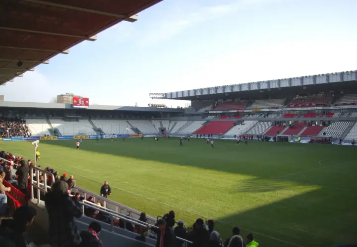 Widok na murawę i trybuny stadionu Cracovii. Na boisku trwa mecz, a trybuny zapełniają się kibicami.