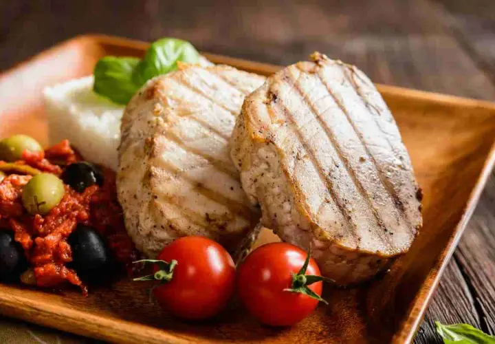 Delizioso tonno alla piastra con contorno di riso, pomodorini, olive e basilico su un piatto di legno.