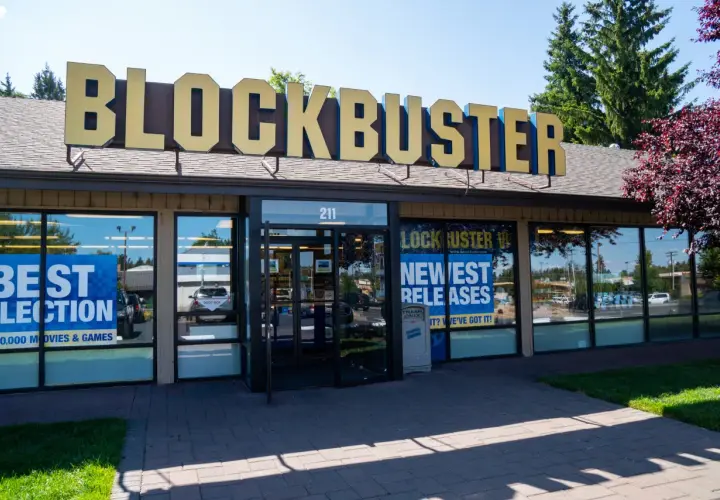 L'insegna dorata del negozio Blockbuster domina l'edificio. Vetrine mostrano "Best Selection" e "Newest Releases", un richiamo nostalgico ai tempi d'oro del blockbuster film.