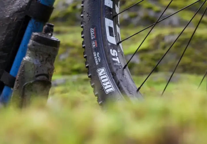 Close-up of a muddy bike tire, showing the "Maxxis Ikon" branding. This tire is ready for a challenging trail review.