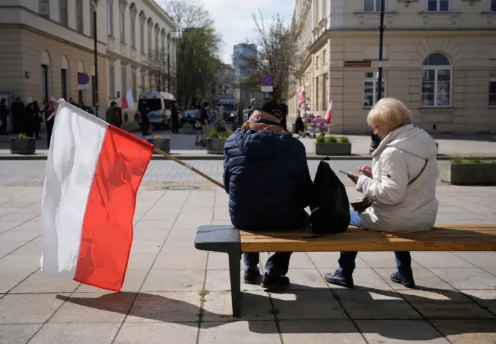 Zwei Personen sitzen auf einer Bank, eine polnische Flagge weht im Wind. Im Hintergrund sind Gebäude und Menschen zu sehen, die an einer Demonstration teilnehmen. Vielleicht geht es um den **ETF Polen**.