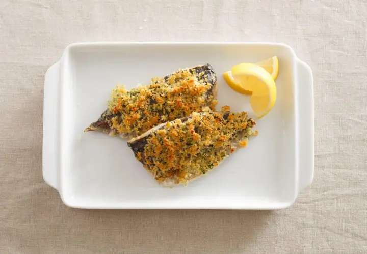 Two fillets of aji fish, baked with a golden breadcrumb topping, served with lemon wedges on a white dish.