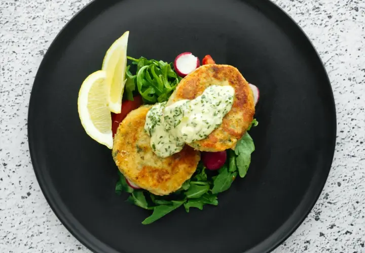 Golden chikuwa tempura patties served with a creamy herb sauce, fresh greens, radishes, and lemon wedges on a black plate.
