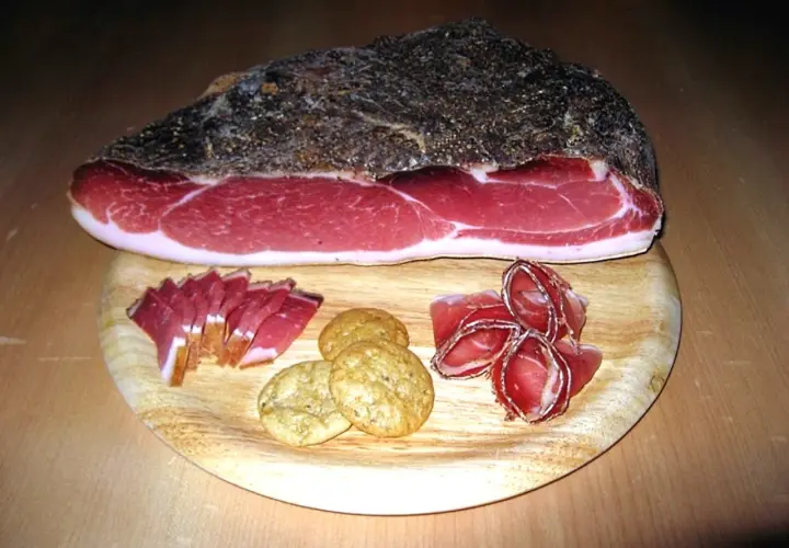 Ein Stück italienischer Speck mit Scheiben und Keksen auf einem Holzbrett.