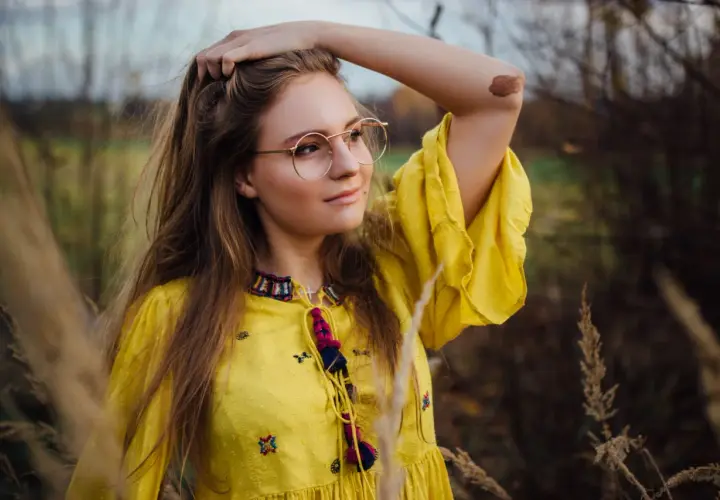Młoda kobieta w okularach i żółtej sukience, uchwycona przez fotografa portretowego, z dłonią we włosach, na tle jesiennej przyrody.
