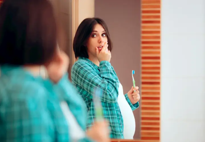 Donna incinta si tocca le labbra, preoccupata per le gengive gonfie in gravidanza. Tiene uno spazzolino, pronta a prendersi cura del suo sorriso.
