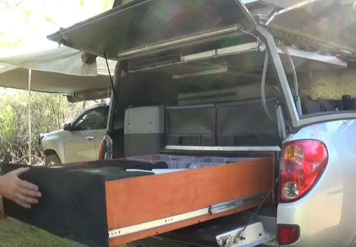 Un homme montre l'intérieur d'une cellule pick-up aménagée avec des tiroirs et des rangements, prête pour l'aventure.