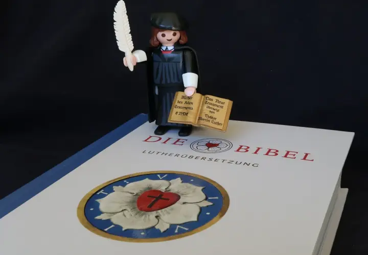 Playmobil-Figur mit Federkiel und Büchern, die den Unterschied zwischen Lutherbibel und katholischer Bibel symbolisiert.