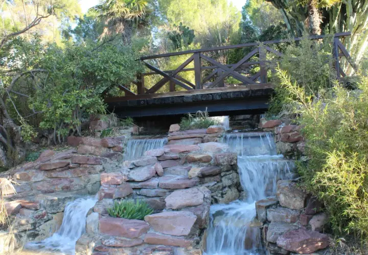 Puente de madera sobre cascadas en uno de los parques de Valencia. Naturaleza exuberante y rocas rojizas crean un paisaje idílico.
