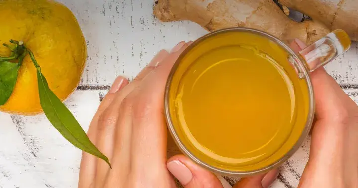 Hände halten ein Glas mit leuchtend gelbem Getränk. Daneben liegen eine Orange und Ingwer. Perfekt, um Kurkuma zuzubereiten.