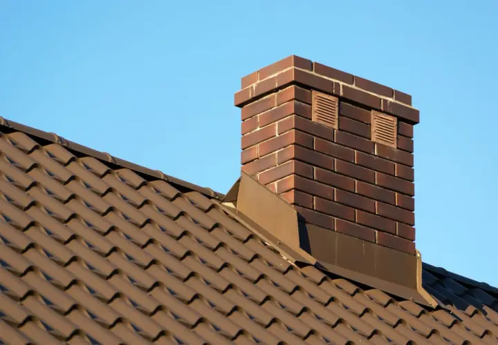 Nowy, brązowy dach z dachówki i wysoki, ceglany komin zewnętrzny ceramiczny z dwoma kratkami wentylacyjnymi na tle błękitnego nieba.