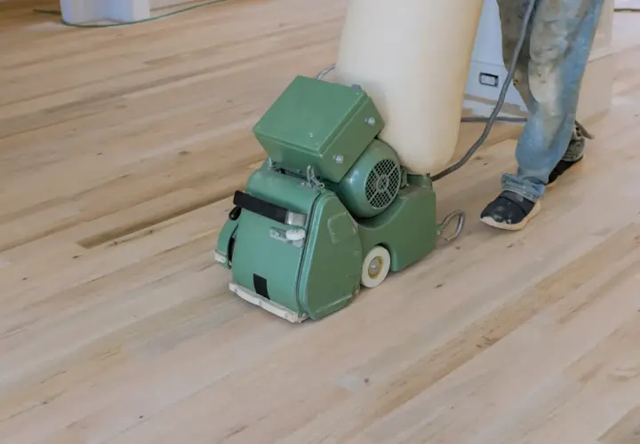 Ein Handwerker schleift mit einer Maschine einen hellen Dielenboden. Staub wird in einem Beutel gesammelt.