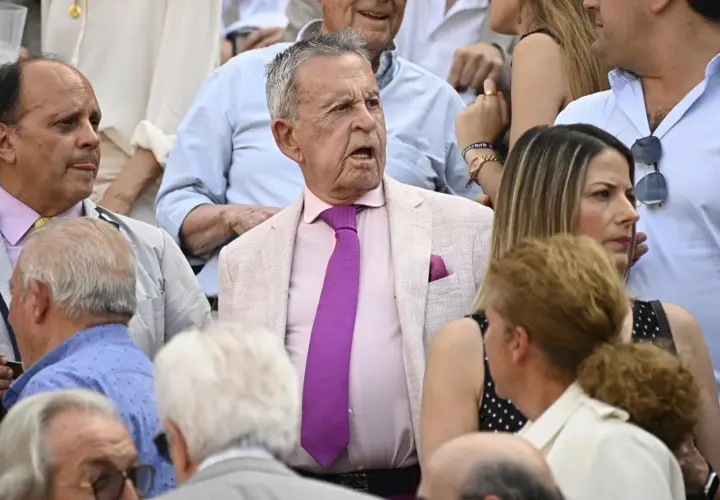 Un grupo de personas, entre ellos algunos famosos en las ventas, observan atentamente un evento. El hombre del centro, con traje rosa y corbata morada, parece sorprendido.
