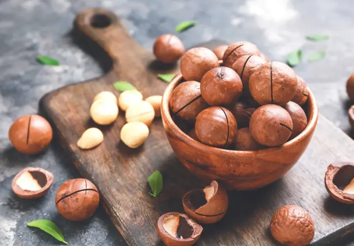 Noci macadamia in una ciotola di legno, con alcune sparse sul tagliere. Scopri i noci macadamia benefici per la tua salute.
