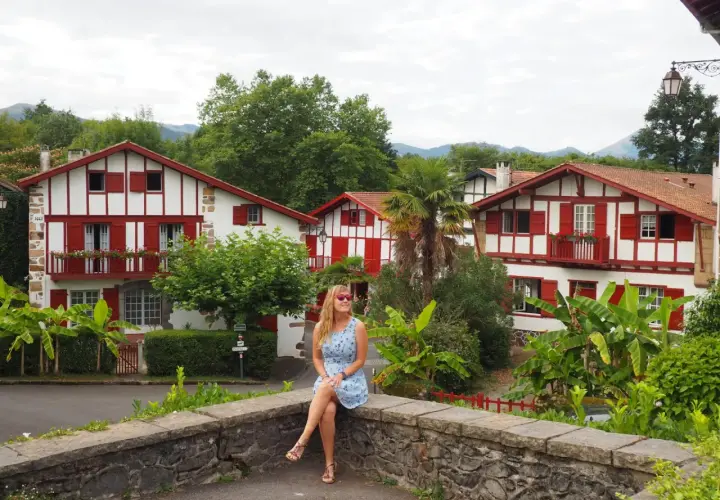Mujer sentada en un muro de piedra, con casas blancas y rojas típicas del Pirineo Vasco al fondo.