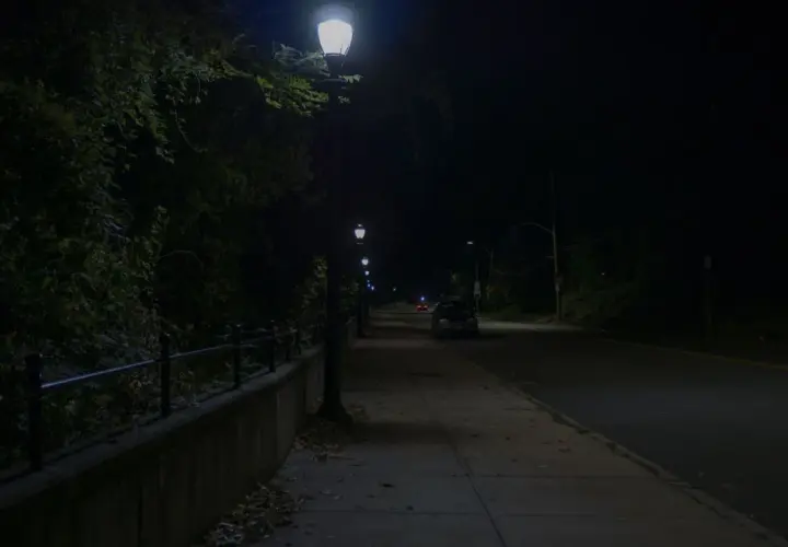 Nocna ulica z latarniami, chodnikiem i samochodami. Ciemność i światło lamp tworzą atmosferę jak z filmu, gdzie nawet kurza ślepota wydaje się możliwa.
