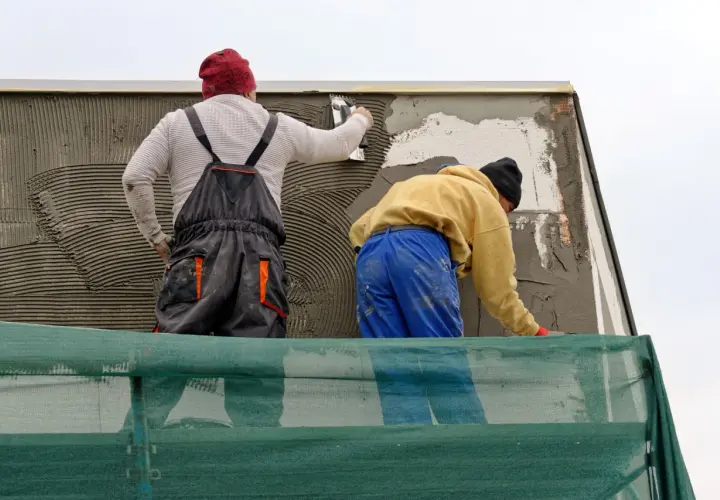 Lavoratori applicano rasatura armata con rete in fibra di vetro su un muro esterno, protetti da un telo verde.