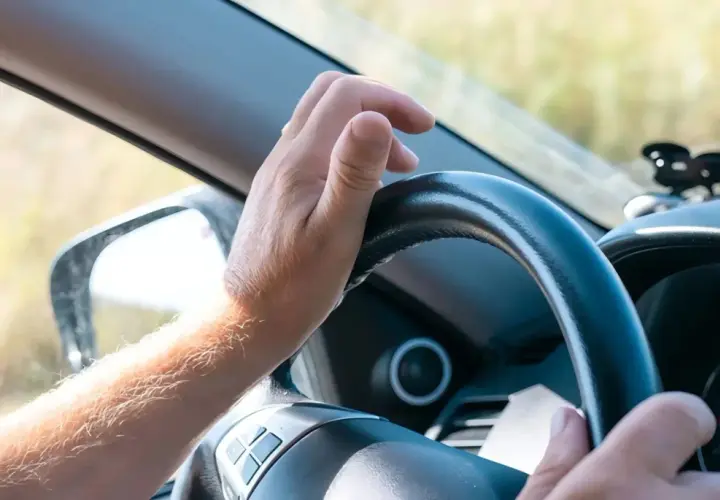 Mano sujetando el volante, con el brazo peludo visible. La dirección del coche dura se siente firme mientras el conductor avanza.