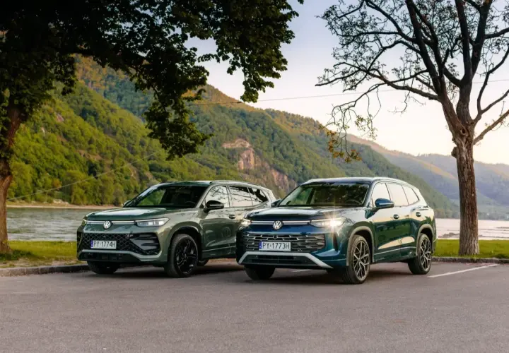 Dwa nowe VW Tayron, zielony i granatowy, zaparkowane nad rzeką, z górami w tle.