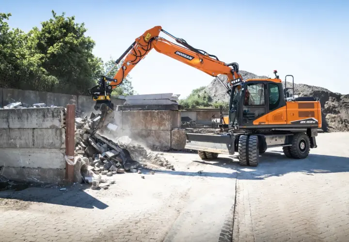Pomarańczowa koparka Doosan DX150W pracuje na placu budowy w Poznaniu, rozbierając betonowe ściany.