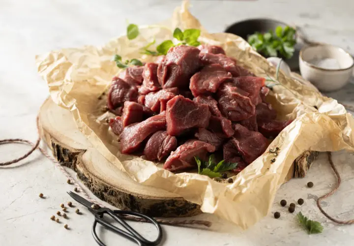 Pokrojone w kostkę mięso dzika, gotowe na aromatyczny gulasz z dzika. Obok gałązki ziół i przyprawy.