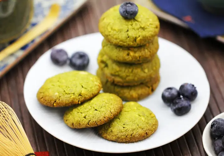 Grüne matcha kekse mit Blaubeeren auf einem Teller, daneben ein Matcha-Schneebesen.
