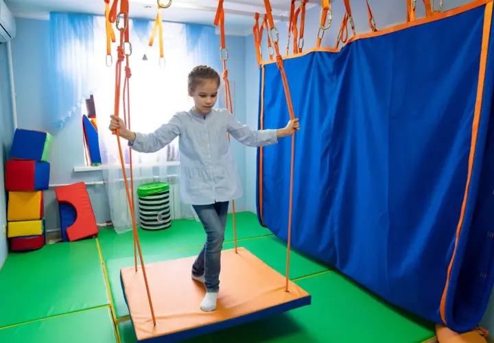 Dziewczynka na huśtawce w sali do terapii sensorycznej. Terapia sensoryczna na czym polega? Na stymulacji zmysłów poprzez ruch i zabawę.