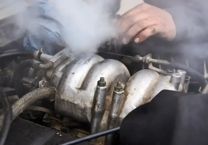 Silnik samochodowy dymi, wydobywa się z niego biały dym. Mechanik próbuje zlokalizować problem, być może z układem paliwowym lub chłodzenia.