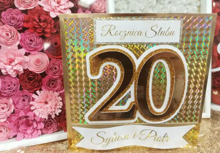 Złota kartka z napisem "Rocznica Ślubu" i liczbą "20" dla Sylwii i Piotra. Obok ramka z różowymi papierowymi kwiatami.